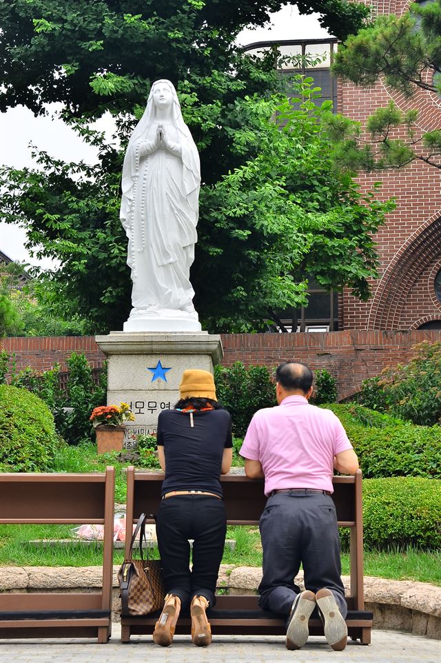 중년의 남녀가 명동성당 뒷동산 성모 마리아상 앞에서 무릎을 꿇고 간절하게 기도하고 있다.