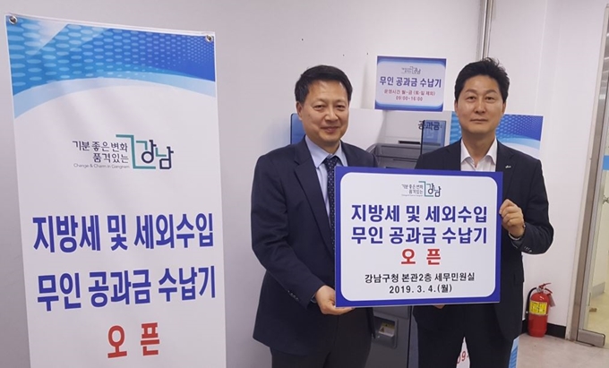 이정헌(왼쪽) 강남구청 세무관리과장이 신한은행 강남구청 지점장과 지난 4일 강남구청 본관 2층 세무과에서 전국 첫 세무과 내 무인 공과금 수납기 설치를 축하하는 팻말을 들고 있다. 강남구 제공