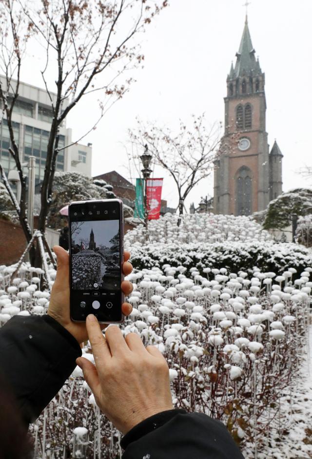 중부지방 곳곳에 눈이 내리는 가운데 13일 오전 서울 중구 명동거리에서 한 시민이 스마트폰으로 눈 덮인 명동성당을 찍고 있다. 눈이 온 뒤 강추위가 닥쳐 서울의 14일 아침 최저기온은 영하 10도, 체감온도는 영하 15도로 예보됐다. 뉴스1