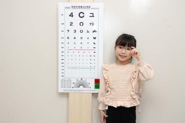 A child wearing glasses stands in front of an eye chart. Courtesy of Kim's Eye Hospital